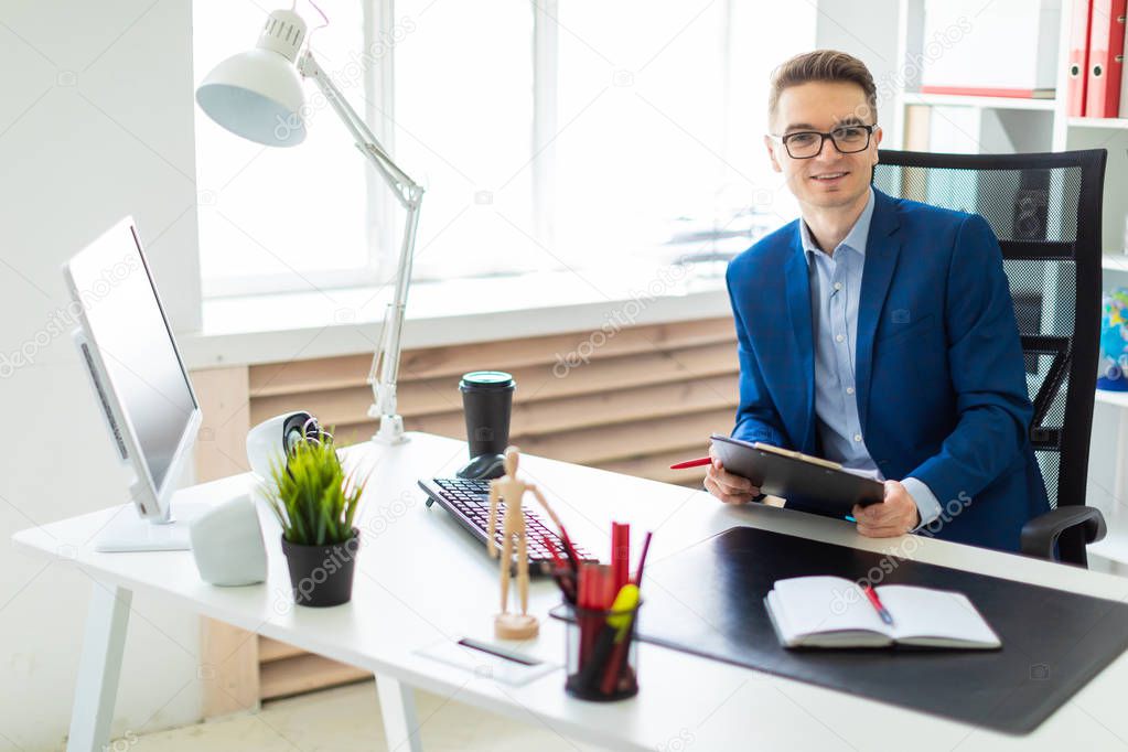 A young man sits at a table in the office and holds documents and a pen in his hands.