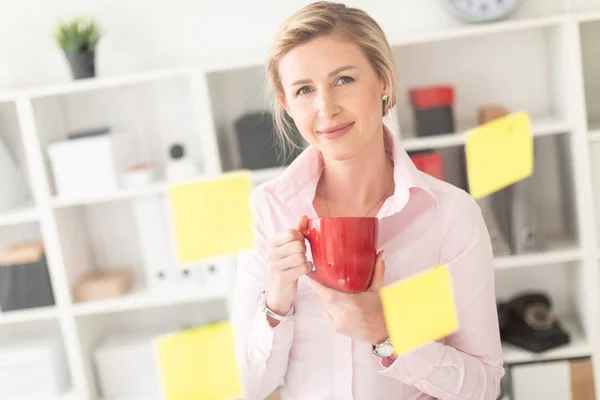 Молодая блондинка стоит в офисе рядом с прозрачной доской с наклейками и держит в руках красную чашку. . — стоковое фото