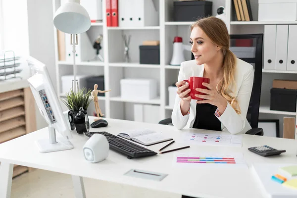 Una joven se sienta en un escritorio de computadora en la oficina y sostiene una taza roja en su mano. Antes de que la chica en la mesa son diagramas . — Foto de Stock
