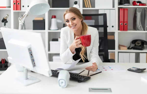 Молодая девушка сидит за компьютерным столом в офисе и держит в руках чашку и документы. Перед девушкой на столе диаграммы . — стоковое фото