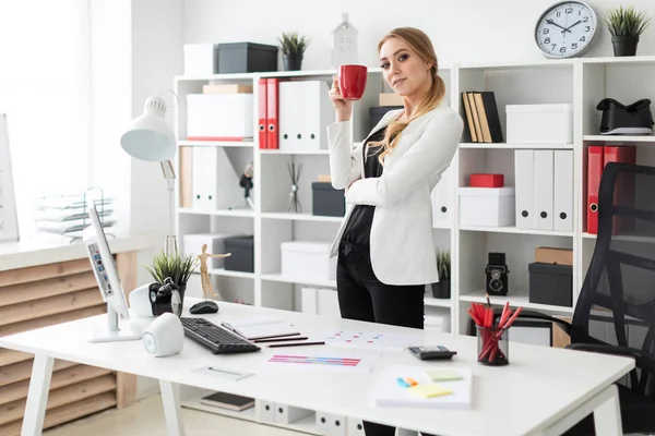 Una joven se para en la oficina cerca de un escritorio de computadora y sostiene una taza roja en sus manos . — Foto de Stock