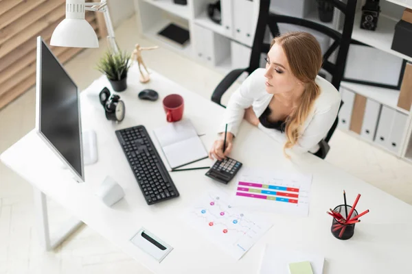 Een jong meisje aan een computerbureau op kantoor zit en rekent op een rekenmachine. Voordat het meisje zijn er documenten. — Stockfoto