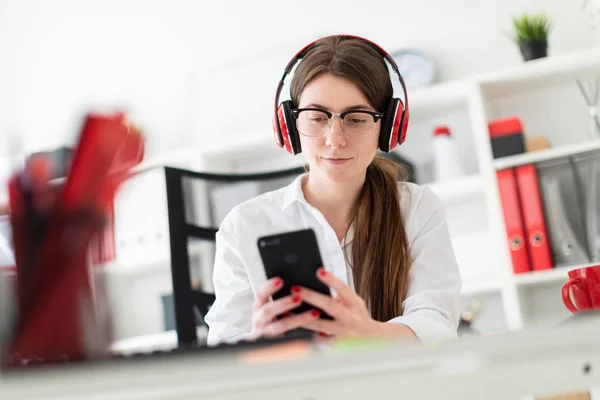 En ung flicka sitter i hörlurar vid ett bord på kontoret och innehar en telefon i handen. — Stockfoto