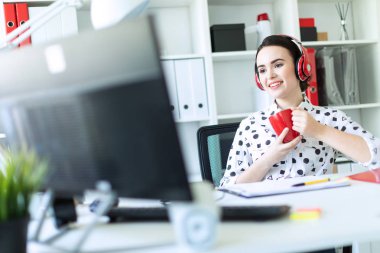 Genç bir kız kulaklık ofiste bir masada oturuyor, kırmızı bir fincan elinde tutan ve ekrana görünüyor.