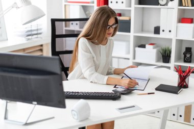 Genç bir kız ofiste, bilgisayar masasında oturuyor onun elinde kalem tutan ve notlar belgede yapma.