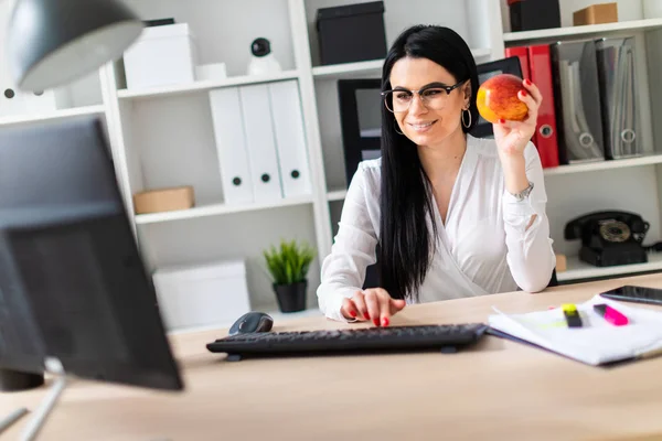 Młoda dziewczyna siedzi przy biurku komputer, w ręku trzyma jabłko i drukuje na klawiaturze. — Zdjęcie stockowe