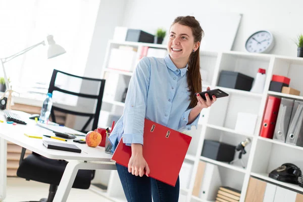 En ung flicka på kontoret är stående, lutad mot ett bord, och håller en telefon och en mapp med dokument. — Stockfoto