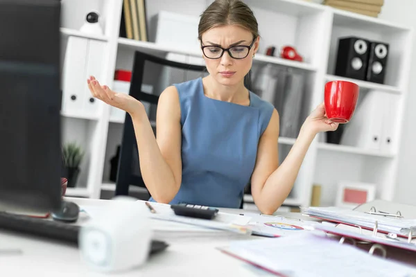 Uma jovem está sentada em uma mesa no escritório, segurando um copo vermelho na mão e trabalhando com documentos. . — Fotografia de Stock