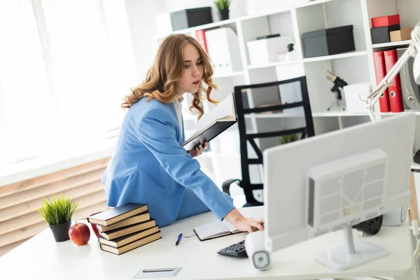 Mooi jong meisje zitten aan de balie in kantoor, bedrijf van een boek in haar hand en kijken naar de monitor. — Stockfoto