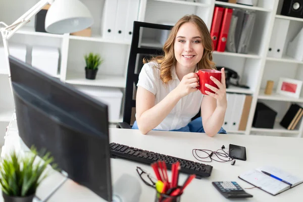 Красивая молодая девушка работает с компьютером в офисе. У девушки в руках красная чашка. . — стоковое фото