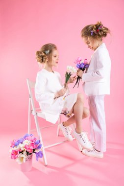 Açık renk elbiseli ikizler çiçek buketleriyle pembe arka planda bir sandalyenin yanında poz veriyorlar..