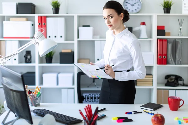 Bir genç kız ofiste masanın yanında duruyor ve onun elinde bir levha notları ve bir kalem tutan.