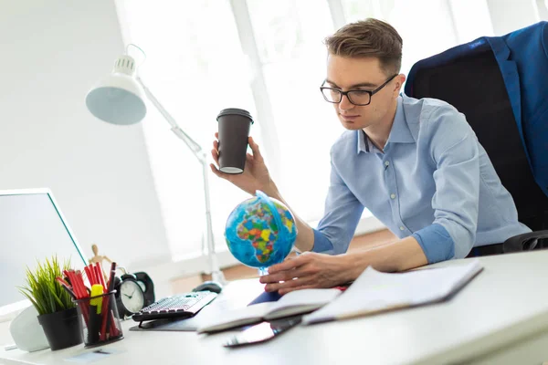 A young man sits in the office at a computer desk and holds a glass of coffee in his hand. A young man faces a globe.
