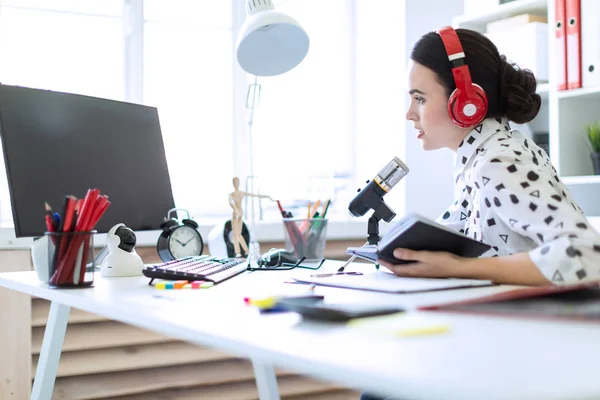 Vacker ung flicka sitter i hörlurar och med ett anteckningsblock vid bordet på kontoret och talar i mikrofonen. — Stockfoto