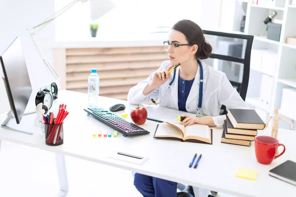 A beautiful young girl in a white robe sits at a table, holds a marker and a pen in her hand and works with a computer and documents.