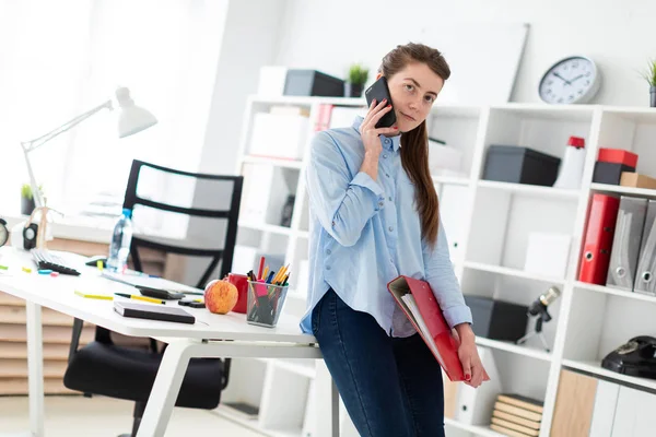 En ung flicka på kontoret är stående, lutad mot ett bord, och håller en telefon och en mapp med dokument. — Stockfoto