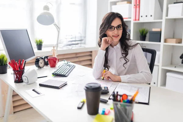 Een jong meisje zit aan de tafel van office en een pen in haar linkerhand houdt. — Stockfoto