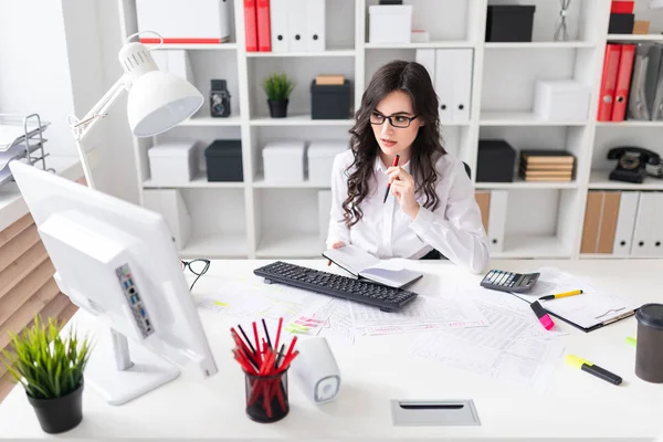 Een jong meisje werkt op de computer in het kantoor en houdt een pen en notitieblok in haar hand. — Stockfoto