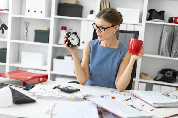 Молодая девушка сидит за столом в своем офисе, держа будильник и красную чашку . — стоковое фото