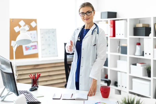 Una joven vestida con una túnica blanca se para cerca del escritorio de la oficina y extiende su mano derecha hacia adelante. Un estetoscopio cuelga alrededor de su cuello . — Foto de Stock