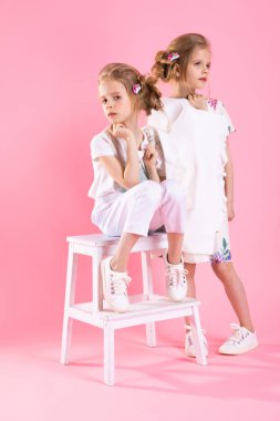 İkiz kızlar parlak giysili iki adım üzerinde pembe bir arka plan ile merdivenlerden yakınındaki poz.
