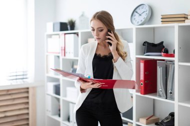 Genç bir kız ofis raf yanında duran, telefonda konuşurken ve belgeler ile bir klasör holding.