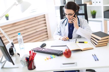 Beyaz bir elbise içinde güzel bir genç kız masada oturmuş, elinde kalem tutan, telefonda konuşurken ve belgeleri seyir.