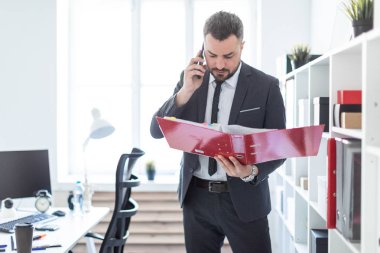 Adam raf ofiste yakın duran, telefonda konuşurken ve bir klasör elinde tutan.