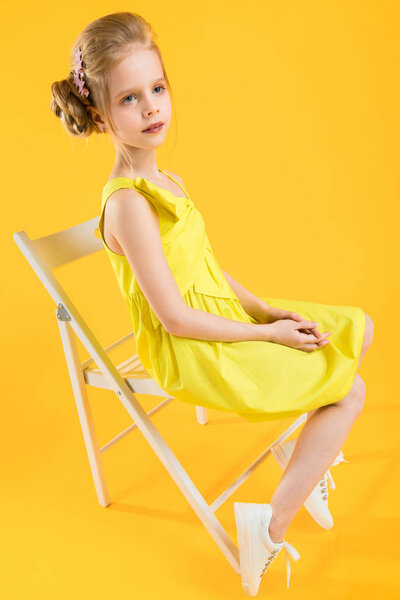 A girl is sitting on a white chair on a yellow background.