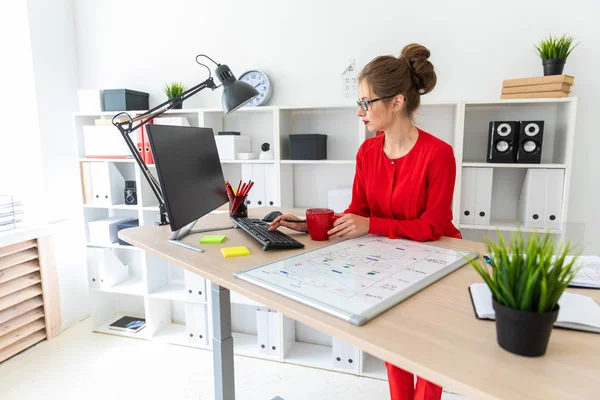 Młoda dziewczyna siedzi przy biurku w biurze, trzyma kubek czerwony w jej dłoni i wpisywanie tekstu na klawiaturze. — Zdjęcie stockowe