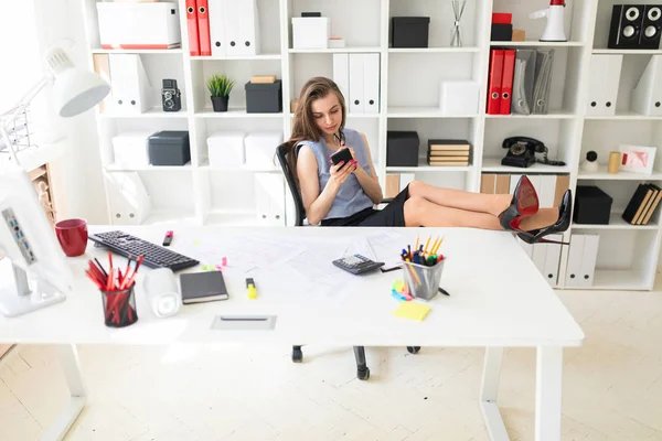 Una hermosa joven en la oficina ha puesto sus pies sobre la mesa y sostiene gafas y un teléfono . —  Fotos de Stock