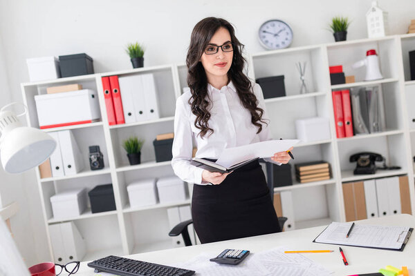 A beautiful young girl stands near an office desk and holds a pen and a notebook in her hands. The girl is negotiating.