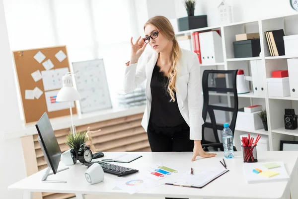 Una joven se para cerca de una mesa en la oficina y endereza sus gafas con la mano . —  Fotos de Stock