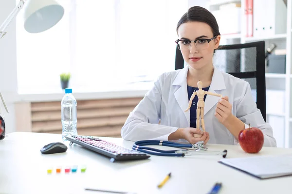 Una hermosa joven con una túnica blanca está sentada en una mesa y sosteniendo una figura de madera de un hombre en sus manos . — Foto de Stock