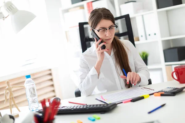 En ung flicka är sitter vid ett bord på kontoret och hålla en penna i handen och prata i telefon. — Stockfoto