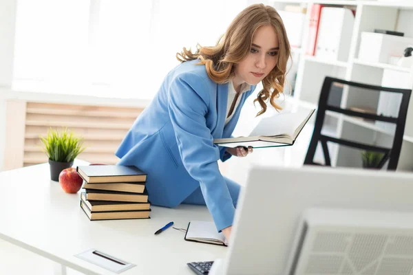 Mooi jong meisje zitten aan de balie in kantoor, bedrijf van een boek in haar hand en kijken naar de monitor. — Stockfoto