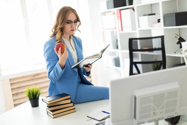 Mooi jong meisje zitten aan de balie in kantoor, bedrijf boek en appel in de hand. — Stockfoto