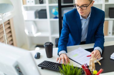 Belgeler ve bir bilgisayar ile ofis ve çalışan bir masada genç bir adam oturuyor.