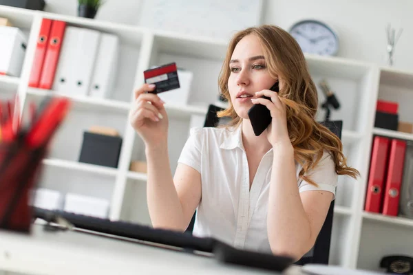 Hermosa joven se sienta en la oficina, tiene una tarjeta bancaria y teléfono en la mano . — Foto de Stock