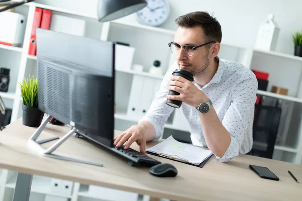 Молодой человек в очках стоит возле стола в офисе, держит в руке стакан кофе и работает с компьютером. — стоковое фото