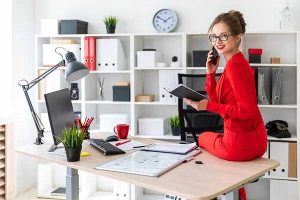 Het jonge meisje ging zitten op de tafel in haar kantoor, gehouden van een notebook in haar handen en was praten over de telefoon. — Stockfoto