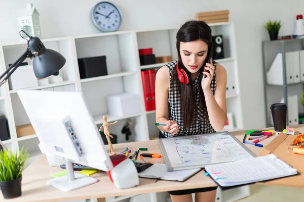 Uma jovem está parada perto de uma mesa, segurando um marcador verde na mão e falando ao telefone. No pescoço, os fones de ouvido das meninas pendurados . — Fotografia de Stock