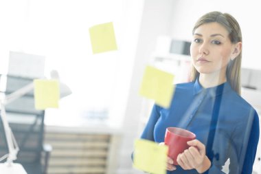 Genç bir kız şeffaf bir yönetim kurulu çıkartmaları ile yakındaki ofiste standları ve kırmızı bir fincan elinde tutan.