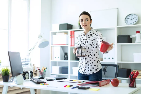 Una joven está de pie en la oficina cerca de la mesa, sosteniendo una tetera en la mano y extendiendo una taza . —  Fotos de Stock