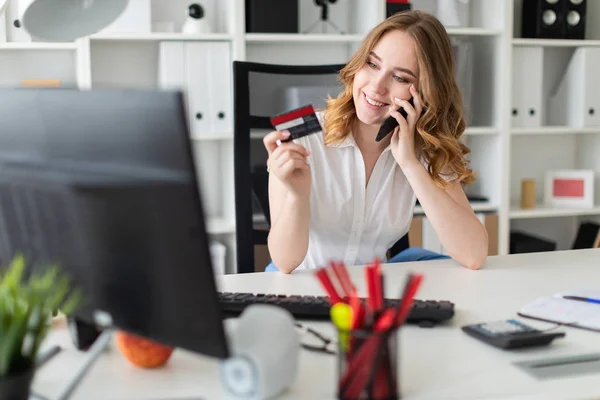 Hermosa joven se sienta en la oficina, tiene una tarjeta bancaria y teléfono en la mano . — Foto de Stock