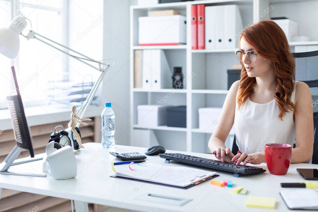 Beautiful young girl is sitting at the desk in the office and typing on the keyboard.