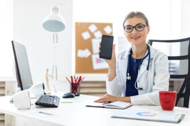 Beyaz bir elbise içinde genç bir kız ofiste bir masada oturur ve bir telefon elinde tutar. Bir stetoskop boynunda asılı.