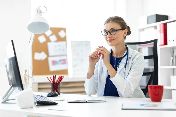 Una giovane ragazza vestita di bianco siede ai tavoli dell'ufficio e tiene in mano una penna. Uno stetoscopio le pende intorno al collo . — Foto Stock