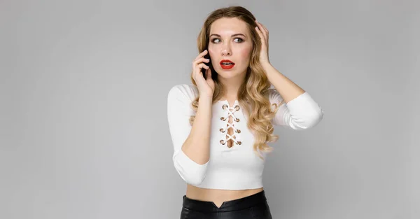 Aantrekkelijke jonge blonde opgewonden zakenvrouw in zwarte en witte kleren glimlachend praten op telefoon staande in kantoor op grijze achtergrond — Stockfoto