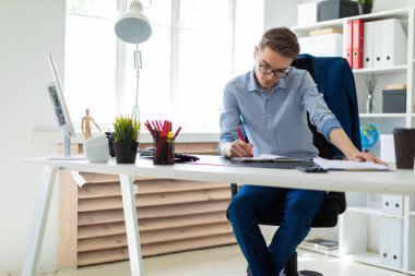 Genç bir adam bir bilgisayar masası ofisinde oturuyor ve bir not defterinde yazıyor.